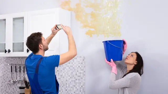 Tenant photographing a ceiling leak while a second person catches drips in a bucket